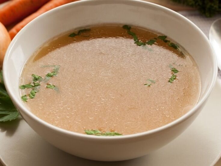 Bowl of clear broth garnished with fresh parsley, surrounded by carrots and an onion, showcasing a healthy and comforting soup ideal for various recipes.