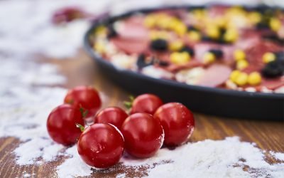 Fresh cherry tomatoes on a wooden surface with a pizza in the background, sprinkled with flour, showcasing ingredients for a delicious homemade meal.